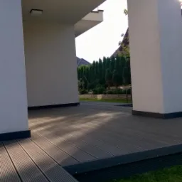 Zadaszony taras z desek kompozytowych w nowoczesnym domu w Krakowie, widok na zadbany ogród z tujami i kamiennym murkiem.