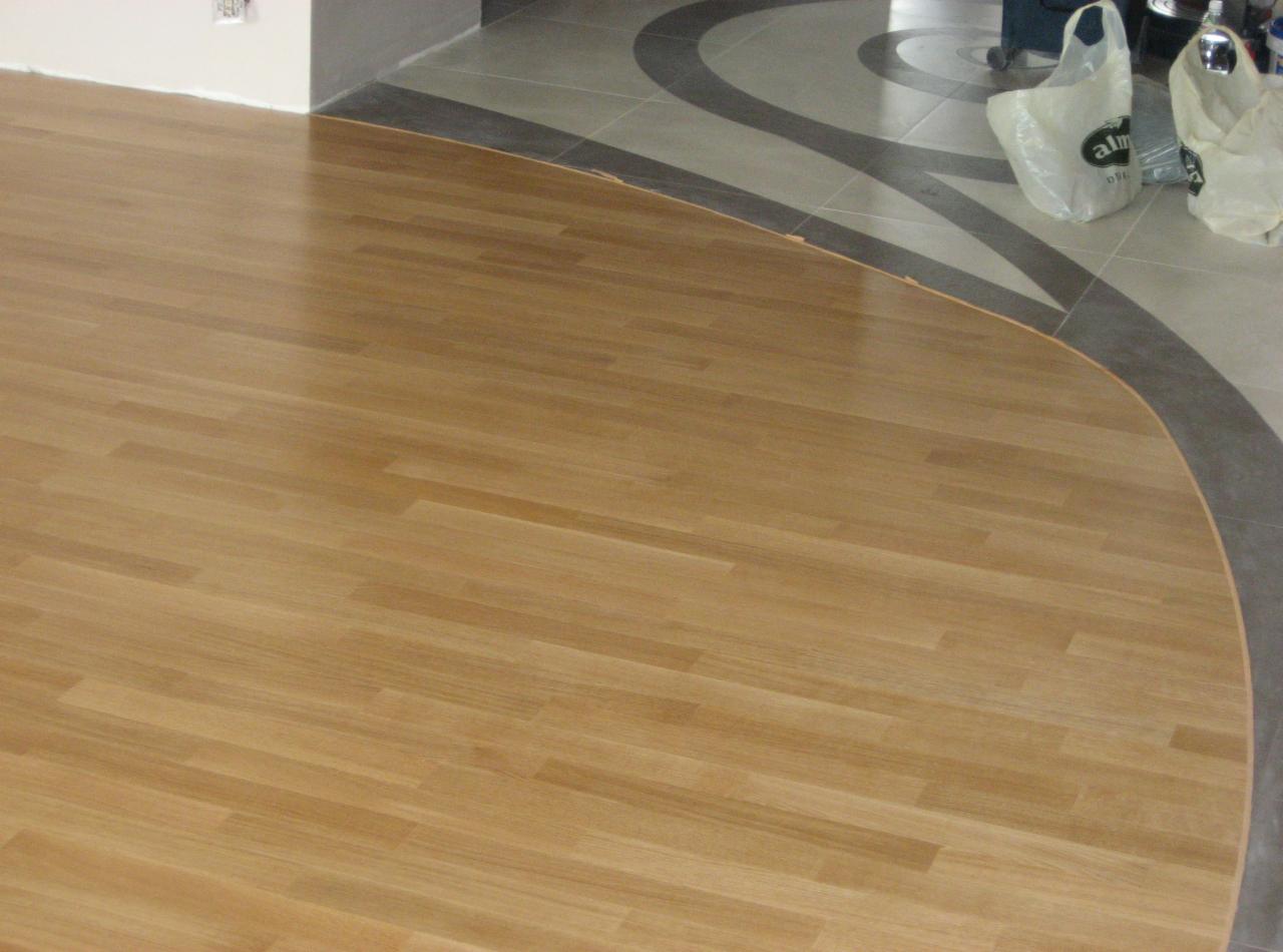 Jasne, drewniane deski parkietu ułożone obok płytek ceramicznych z dekoracyjnym wzorem, widoczne torby foliowe w tle.