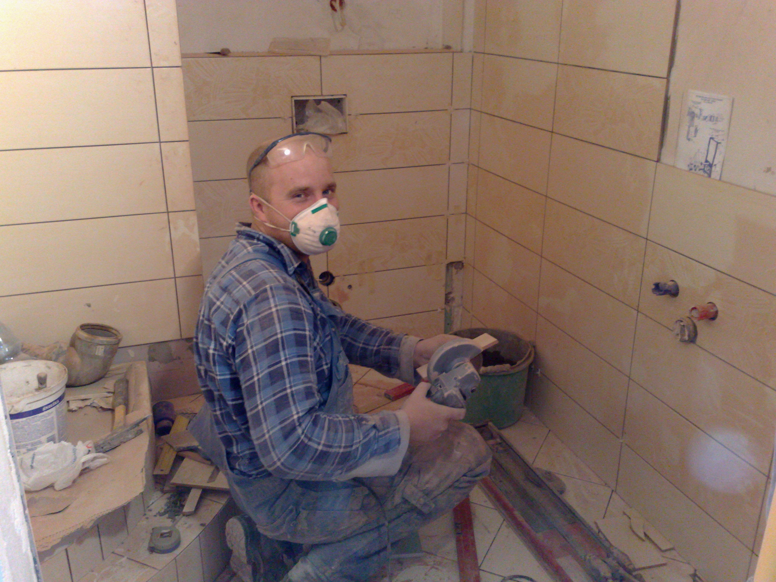 Mężczyzna w masce i okularach ochronnych, klęczący w trakcie docinania płytki ceramicznej szlifierką kątową w łazience z częściowo wyłożonymi kaflami.