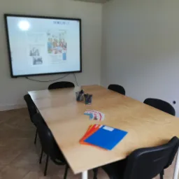 Sala lekcyjna z drewnianym stołem, czarnymi krzesłami i interaktywną tablicą wyświetlającą materiały edukacyjne, w tym zdjęcia grupowe i tekst w języku niemieckim.