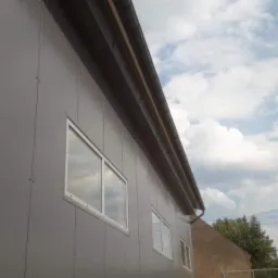 Szara hala z płyt warstwowych z białymi oknami i brązowym orynnowaniem, sfotografowana pod kątem, z widocznym fragmentem nieba i budynkiem w tle.