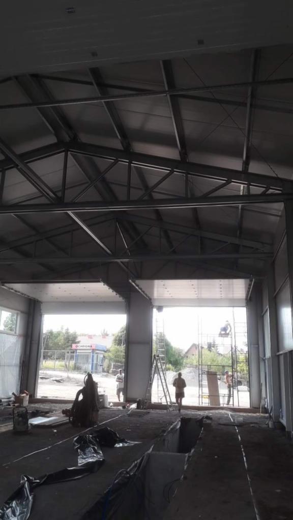 Wnętrze hali w trakcie budowy, widoczne stalowe elementy konstrukcyjne dachu, częściowo zamontowane płyty warstwowe na ścianach, pracownicy w oddali i sprzęt budowlany na podłodze.