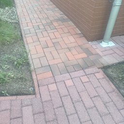 KATARZYNA KONTEK Adamex - Czysta, czerwono-brązowa kostka brukowa ułożona wzdłuż ceglanej ściany budynku z rynną. Wiadro w rogu kadru. Widoczne fragmenty trawy i ziemi.