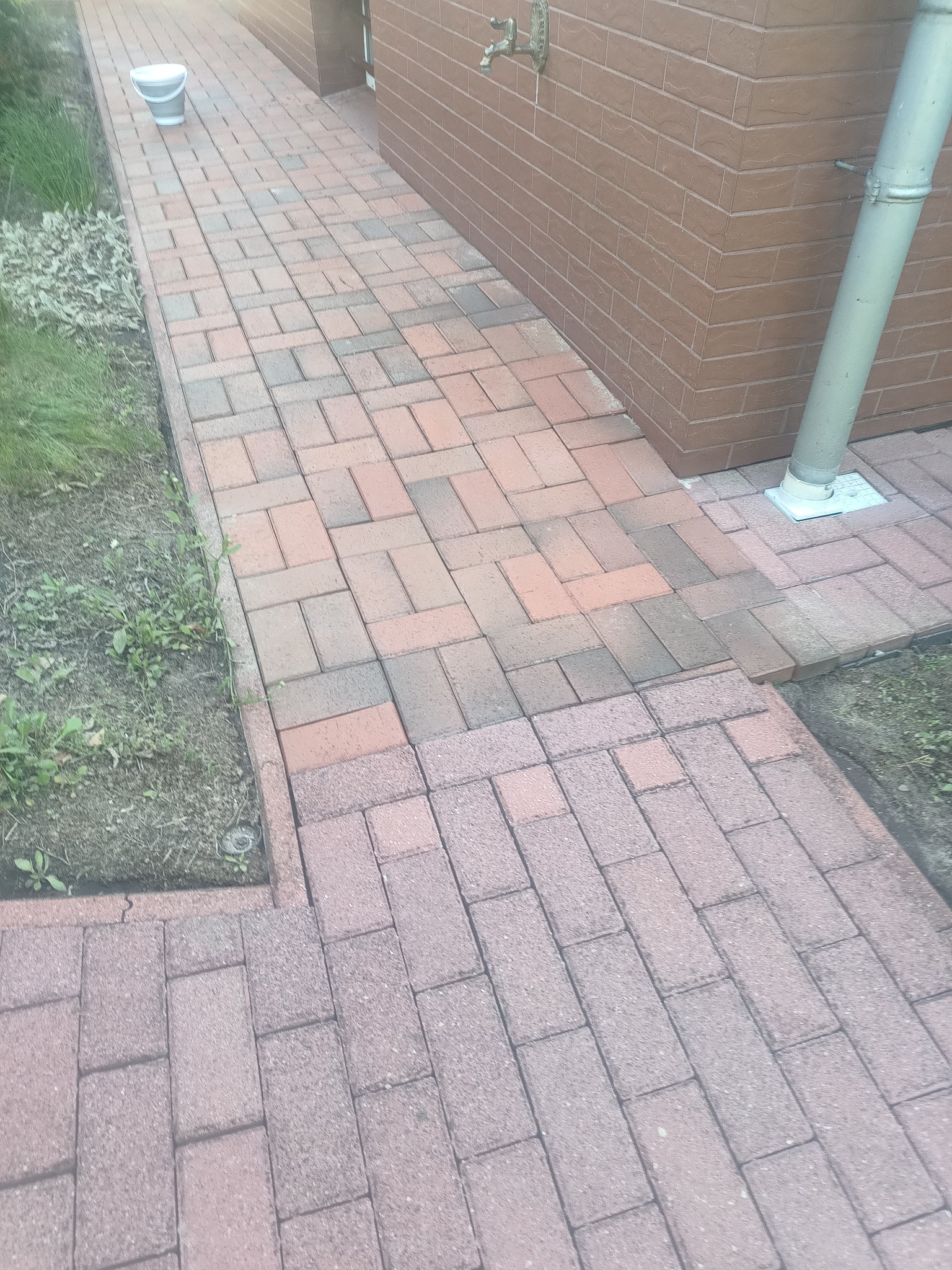 Czysta, czerwono-brązowa kostka brukowa ułożona wzdłuż ceglanej ściany budynku z rynną. Wiadro w rogu kadru. Widoczne fragmenty trawy i ziemi.