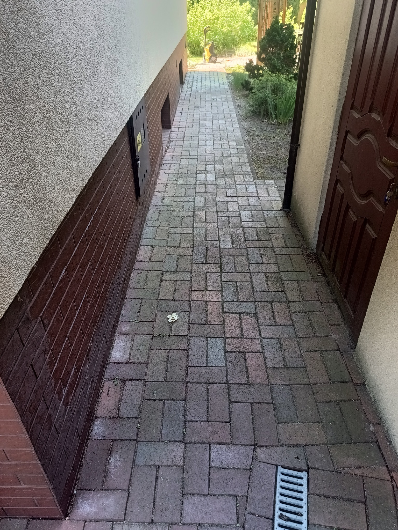 Umyta kostka brukowa na wąskim przejściu między budynkami. Widoczny odpływ liniowy i ślady wilgoci. W tle urządzenie wysokociśnieniowe.