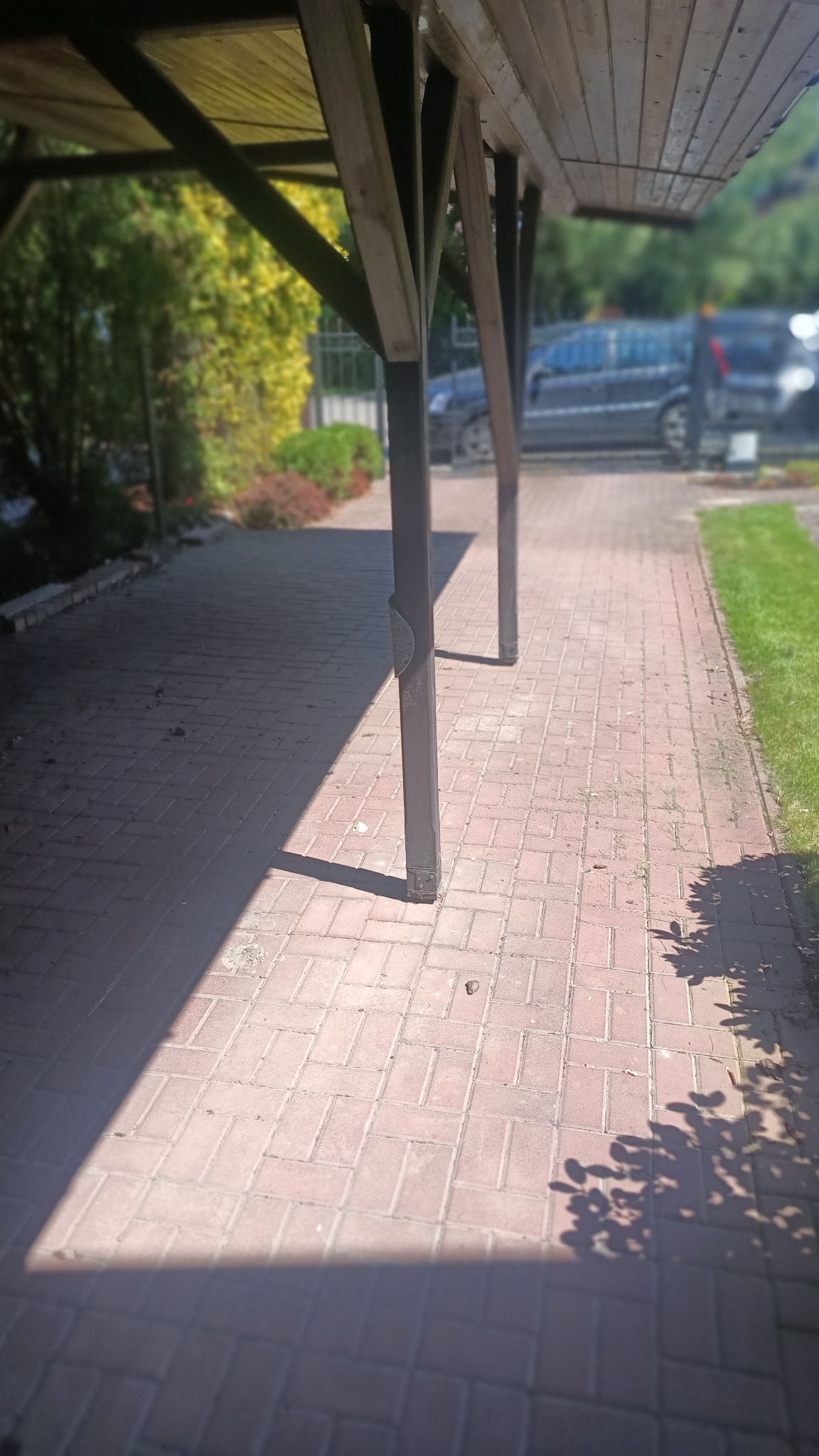 Czerwona kostka brukowa pod zadaszeniem, oświetlona słońcem, z widocznymi cieniami słupów. W tle zieleń i zaparkowany samochód.