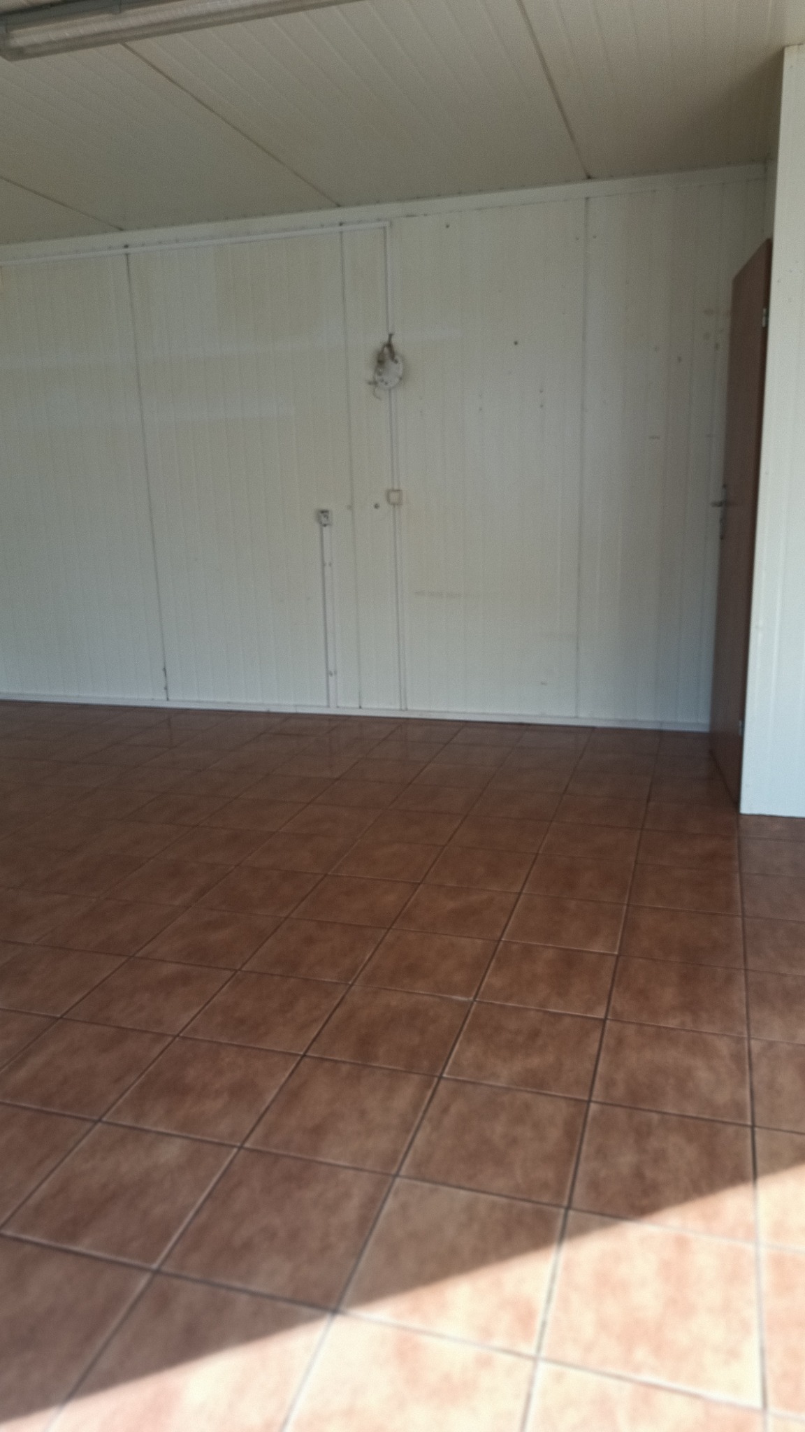 Puste pomieszczenie z brązowymi płytkami na podłodze i panelami ściennymi. Widoczne niedoskonałości na ścianach i suficie, oświetlenie sufitowe i cień na podłodze.