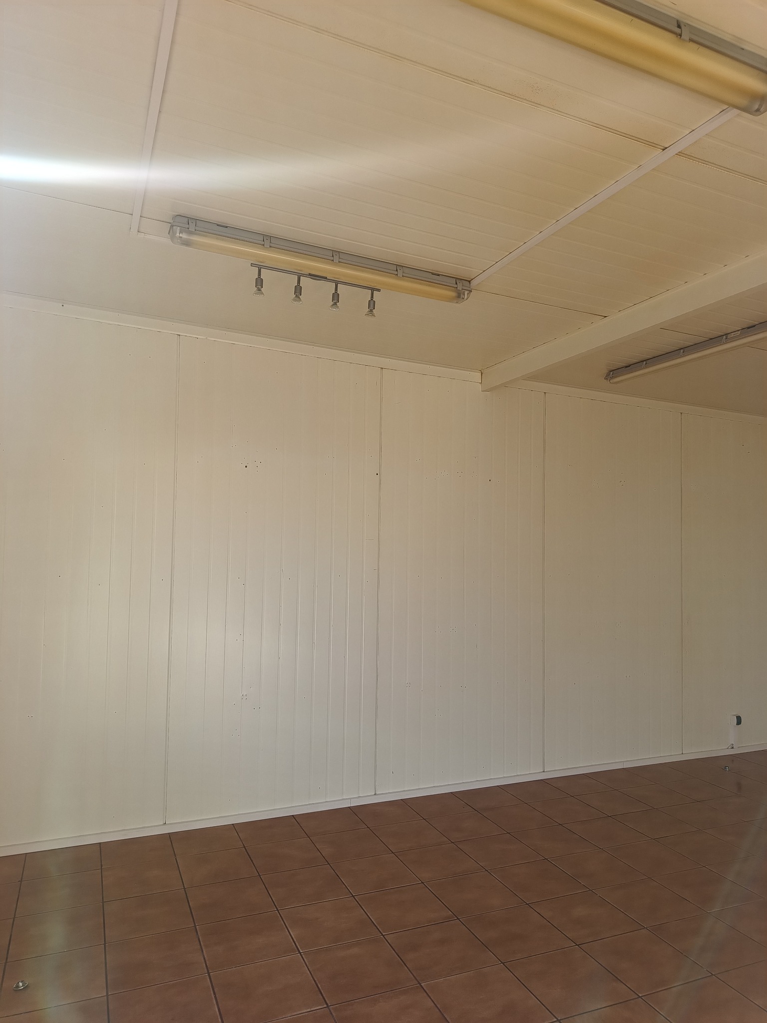 Puste pomieszczenie z brązowymi płytkami podłogowymi i kremowymi panelami ściennymi pod fluorescencyjnym oświetleniem. Widoczny fragment ściany i sufitu.