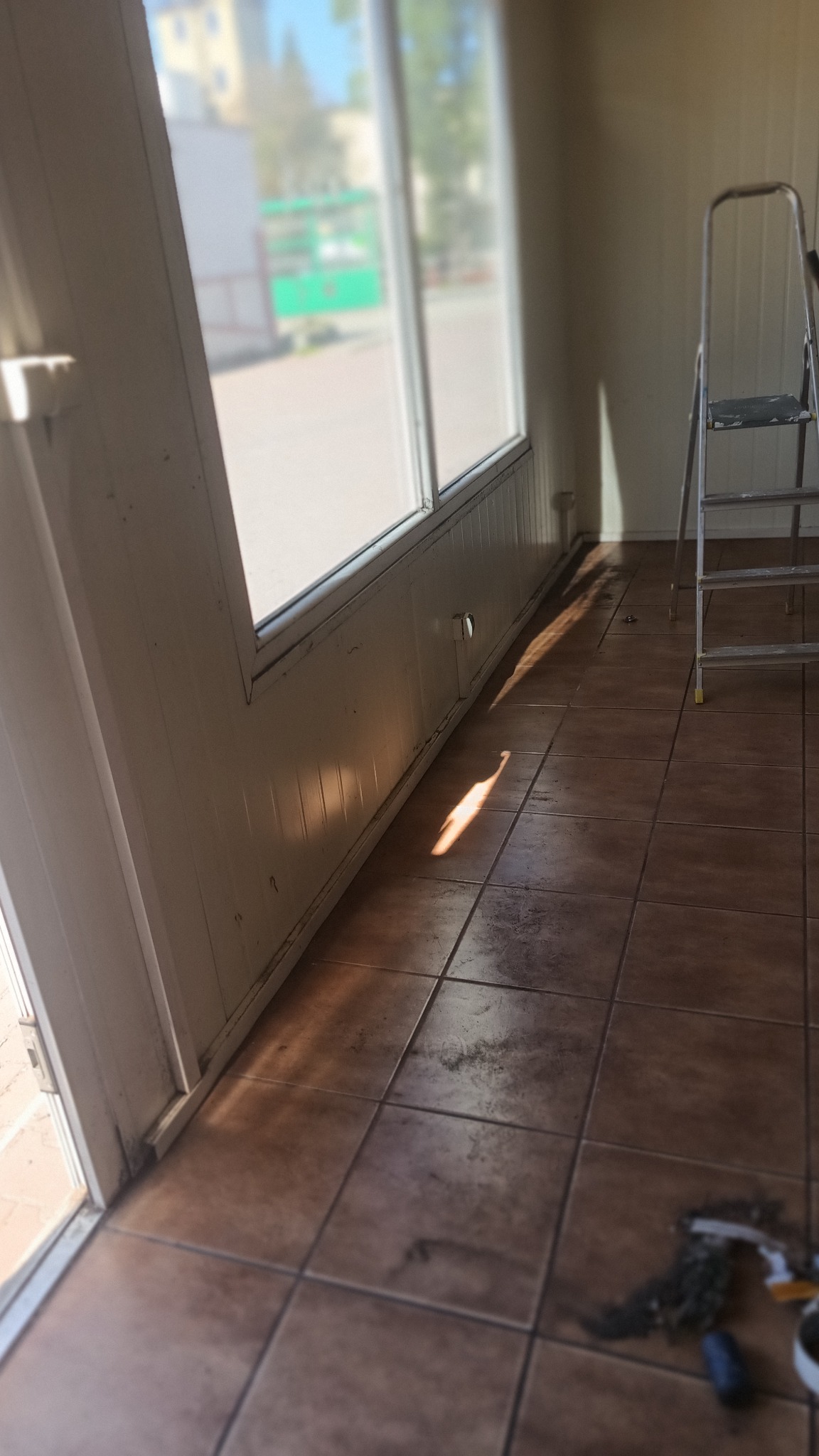Wnętrze z brudną podłogą z płytek i drabiną. Widoczne okno z zanieczyszczeniami. Na podłodze resztki po sprzątaniu, w rogu śmieci. Słoneczny blask podkreśla zabrudzenia.