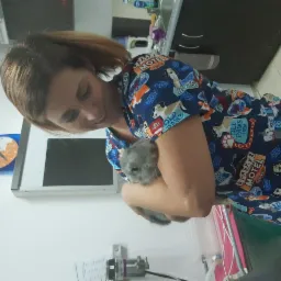 Kobieta w koszuli z motywem zwierzęcym trzyma szynszylę na rękach w gabinecie z zegarem na ścianie i sprzętem medycznym w tle.