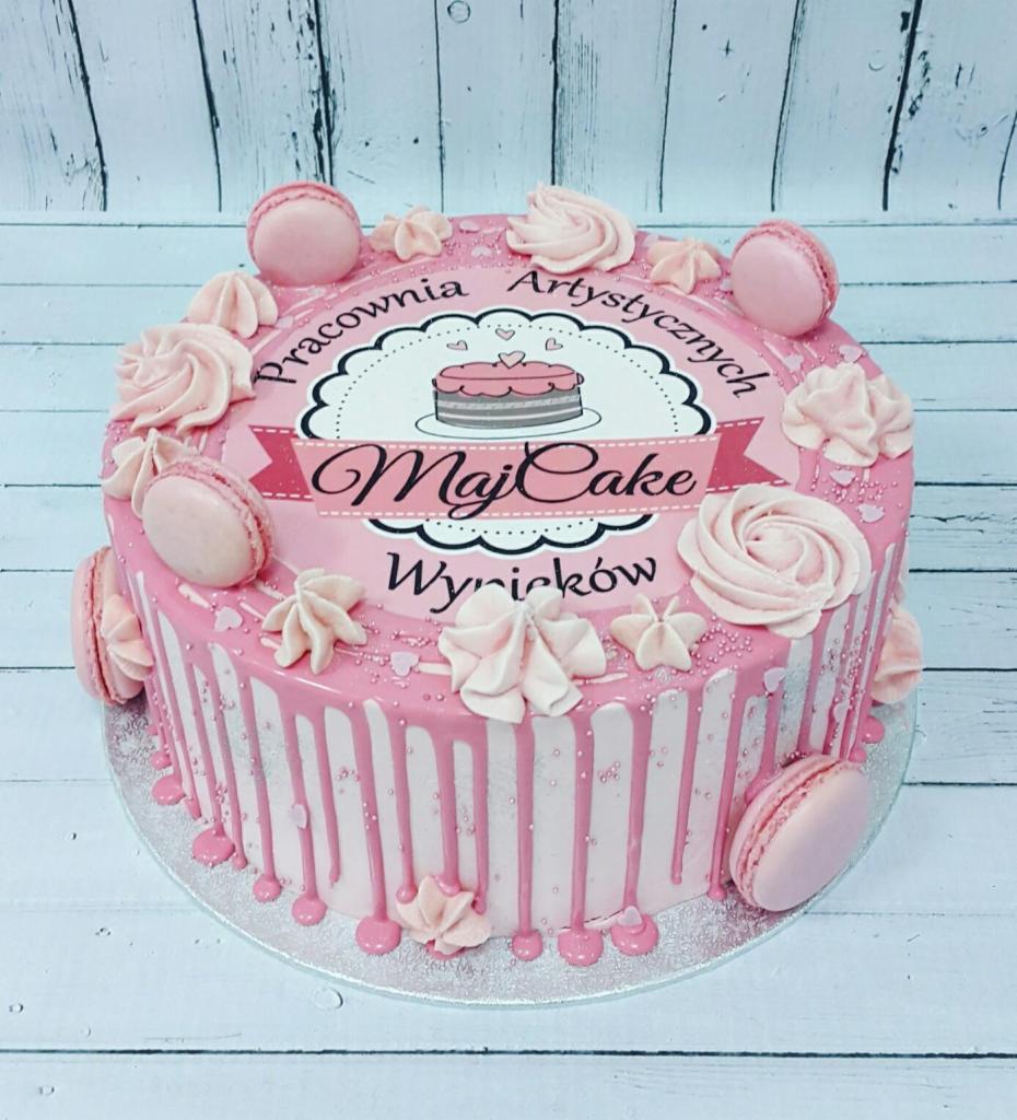 Różowy tort z makaronikami i dekoracjami z kremu, z nadrukiem logo 'Pracownia Artystycznych Wypieków MajCake' na białym drewnianym tle.