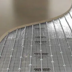 Montaż ogrzewania podłogowego na folii aluminiowej z siatką, widoczne rurki grzewcze i fragment wylewki jastrychowej.