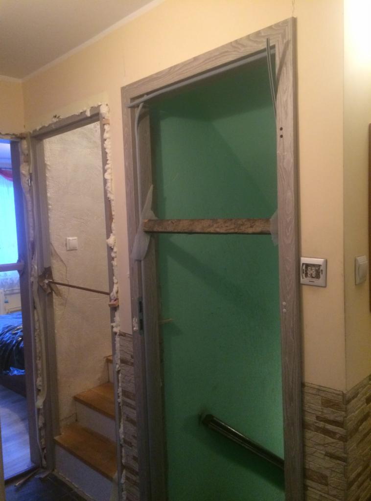 Montaż ościeżnicy drzwi wewnętrznych z użyciem pianki montażowej i rozpórki drewnianej; widoczna surowa ściana i schody w tle.