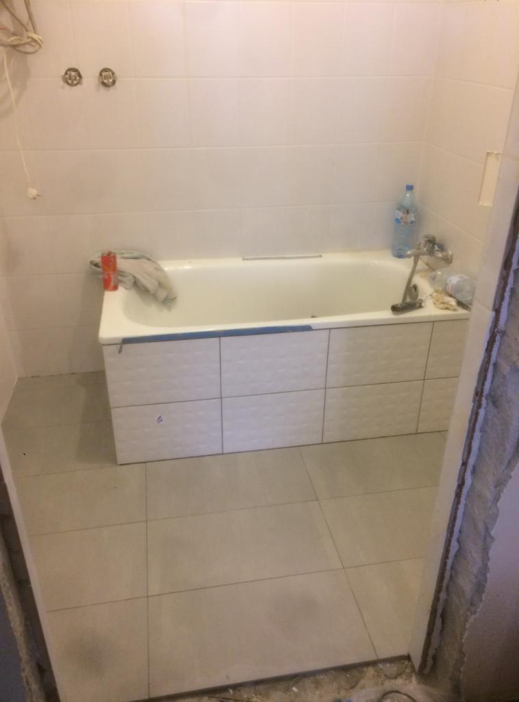 Biała wanna obudowana płytkami z fakturą, świeżo ułożone jasne płytki podłogowe, widoczne niedokończone wykończenie ościeżnicy, butelka wody i puszka na brzegu wanny.