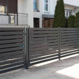 Nowoczesne, poziome, szare ogrodzenie z metalowych profili na tle jasnego domu z balkonem i zielenią.