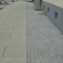 Ułożona nawierzchnia z kostki granitowej w odcieniach szarości, sąsiadująca z chodnikiem z dużych, jasnych płyt betonowych wzdłuż ściany budynku z małymi okienkami wentylacyjnymi.