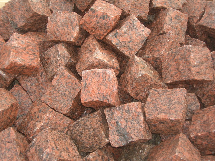 Góra czerwono-brązowych, surowych kostek granitowych o nieregularnej fakturze, z widocznymi ciemnymi plamkami, ułożonych chaotycznie.