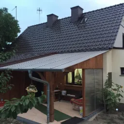 Dom z brązowym dachem z dachówki, drewnianą werandą z przezroczystym zadaszeniem i rynną, widoczne kominy i anteny na dachu.