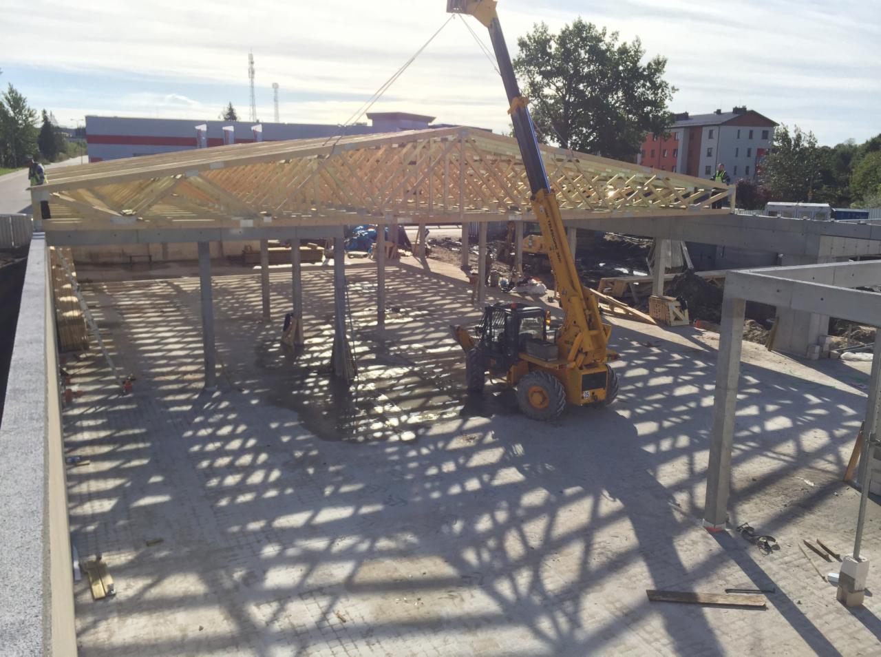 Montaż drewnianej więźby dachowej na betonowych słupach z użyciem dźwigu teleskopowego na placu budowy w słoneczny dzień.