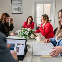 Zespół biura rachunkowego przy pracy: przeglądanie dokumentów, praca na laptopie i tablecie, w tle dekoracja kwiatowa i grafiki na ścianie.