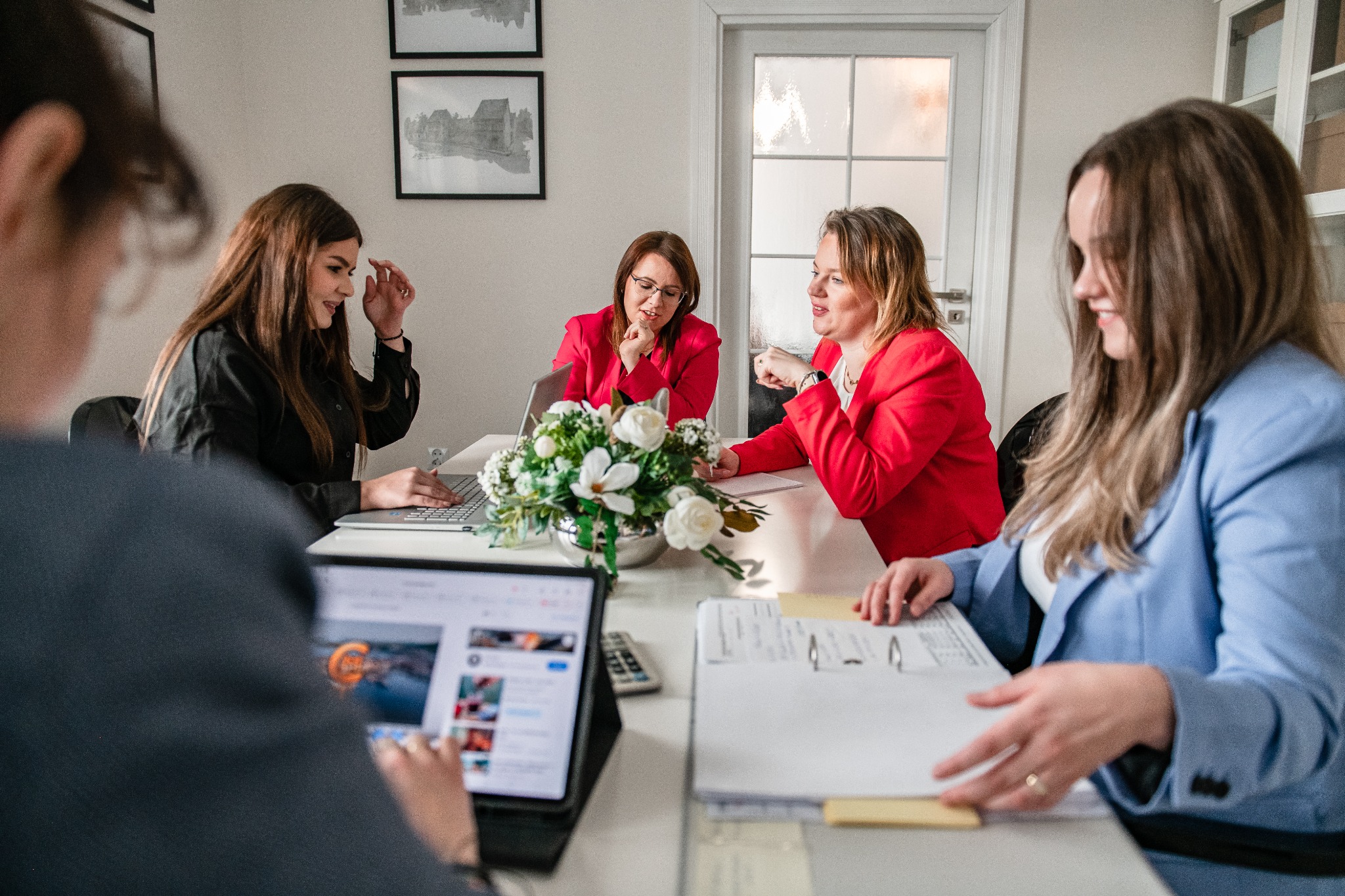 Zespół biura rachunkowego przy pracy: przeglądanie dokumentów, praca na laptopie i tablecie, w tle dekoracja kwiatowa i grafiki na ścianie.