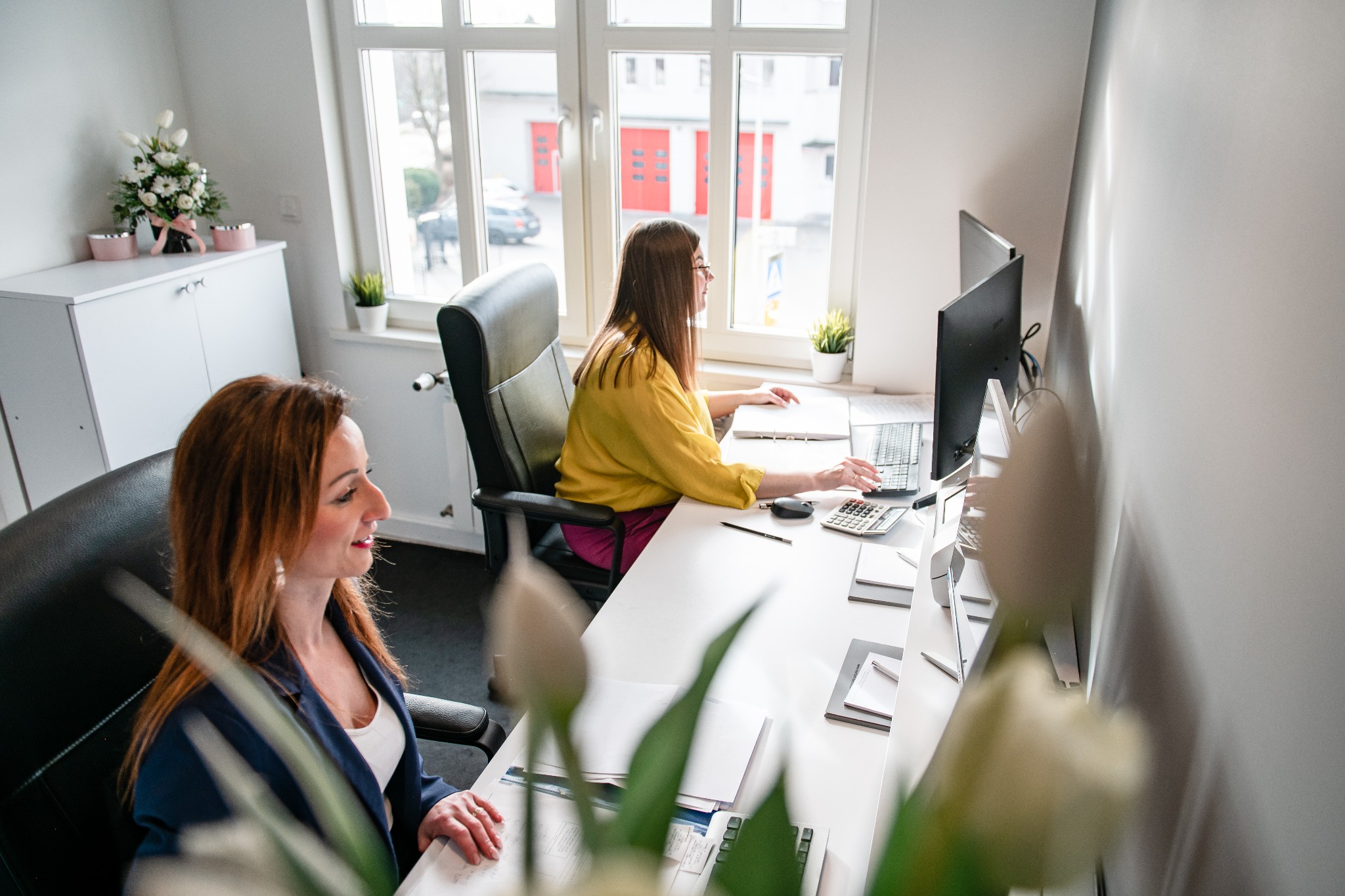 Dwie kobiety w biurze przy białych biurkach, pracujące na komputerach. Widoczne dokumenty, kalkulatory i kwiaty. Jasne wnętrze z dużym oknem w tle.