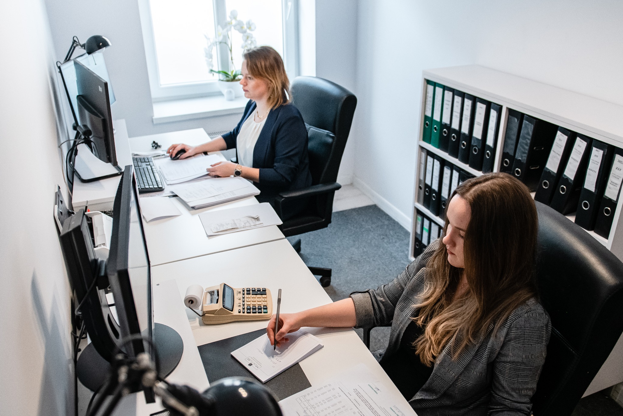 Dwie kobiety pracują w biurze. Jedna przy komputerze, druga pisze notatki obok kalkulatora. W tle regał z segregatorami.