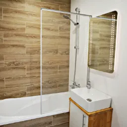 Nowoczesna łazienka po remoncie z wanną za szklanym parawanem, umywalką nablatową na drewnianej szafce i podświetlanym lustrem.