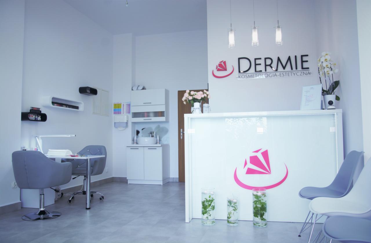 Jasne wnętrze gabinetu kosmetycznego z recepcją, stanowiskiem do manicure z szarymi krzesłami i półkami z lakierami, logo firmy na ścianie i dekoracyjne szklanki z roślinami.