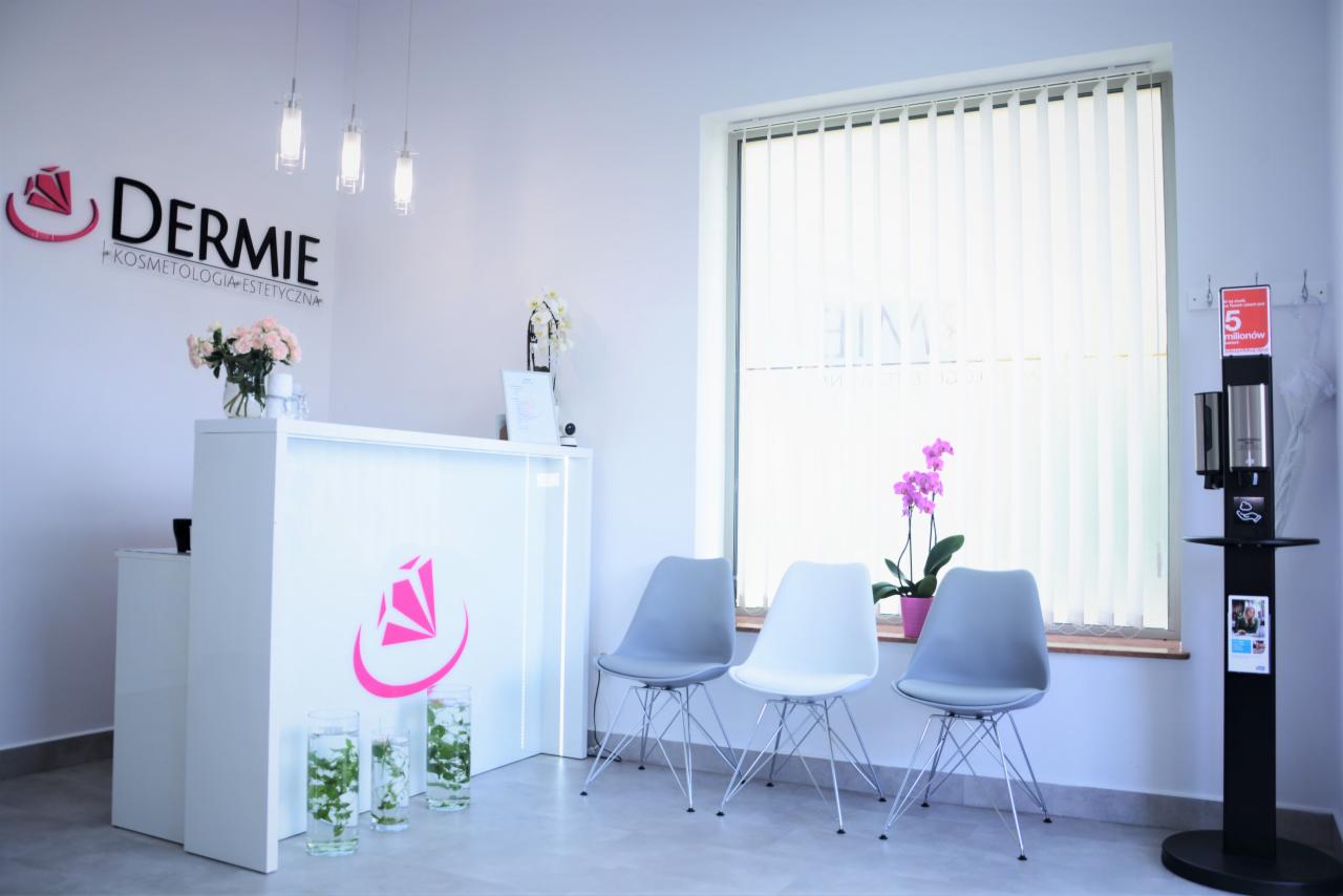 Jasne wnętrze recepcji salonu z logo 'DERMIE Kosmetologia Estetyczna', kwiatami, trzema krzesłami i dozownikiem do dezynfekcji przy oknie.