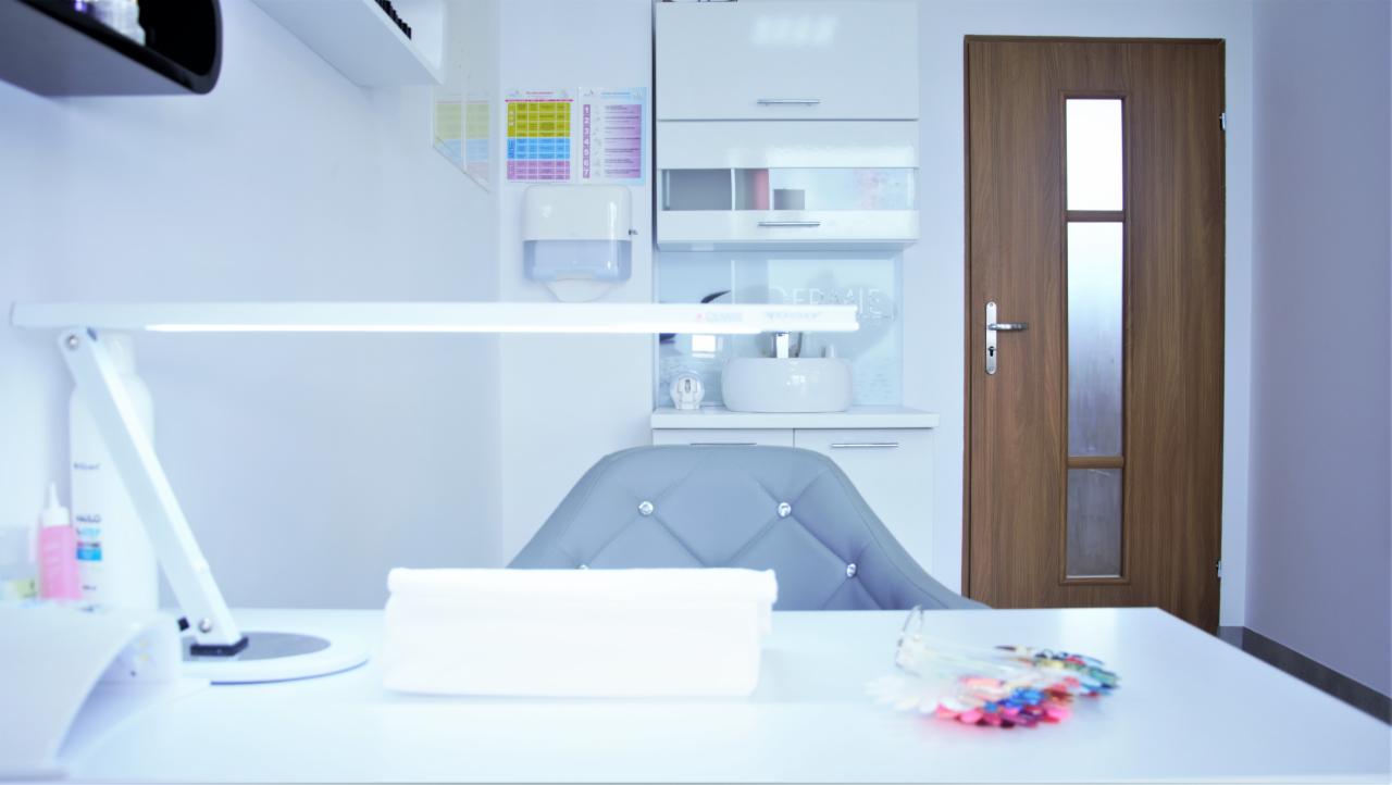 Wnętrze gabinetu manicure z białym biurkiem, lampą LED, paletą kolorów lakierów i szarym krzesłem z kryształkami na tle białych szafek i drewnianych drzwi.