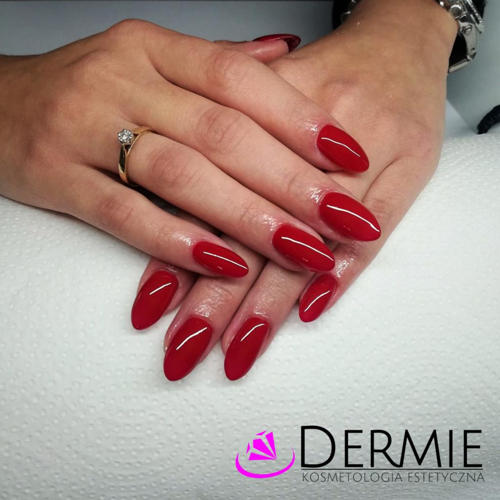 Dłonie z czerwonymi, błyszczącymi paznokciami w kształcie migdała, na palcu złoty pierścionek z brylantem, na dole logo salonu kosmetycznego.