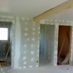 Wnętrze w trakcie prac wykończeniowych: surowe ściany z widocznymi łatami gipsowymi, drewniana belka stropowa, drabina aluminiowa i narzędzia w tle, widoczne otwory drzwiowe.