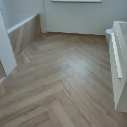 Jasne panele ułożone w jodełkę w wykończonej łazience, widoczna wanna obudowana panelami i fragment białej umywalki