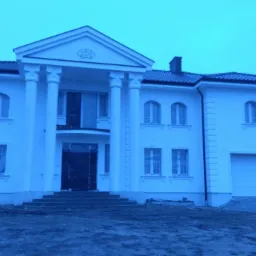 Dwupiętrowy dom z białymi ścianami i kolumnami przed wejściem, dachówka w kolorze grafitowym, okna w kształcie łuku, teren wokół domu niezagospodarowany.