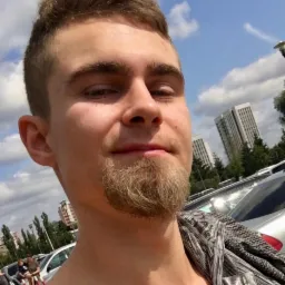 Portret młodego mężczyzny z kozia bródką na tle parkingu w Poznaniu, z budynkami w oddali i błękitnym niebem z chmurami.