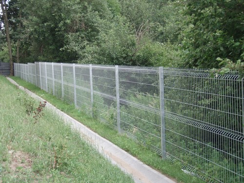 Długi, srebrny panelowy płot z siatki metalowej, rozciągający się wzdłuż betonowego korytka na zboczu porośniętym trawą, z gęstym zadrzewieniem w tle.