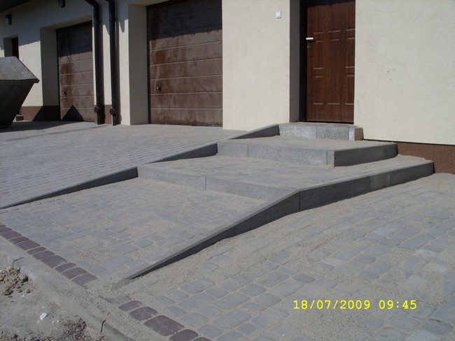 Nowo ułożona nawierzchnia z szarej kostki brukowej przed domem, z rampą i schodami wejściowymi, zasypana piaskiem.