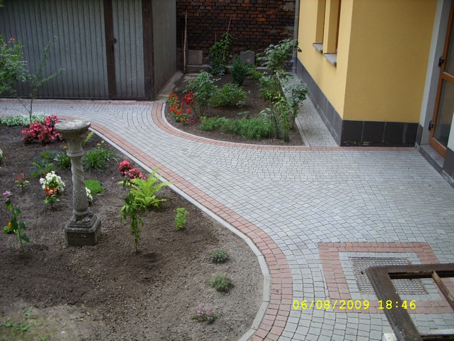 Ścieżka z szarej kostki brukowej z czerwoną obwódką wijąca się przez ogród z kwiatami i rzeźbą, prowadząca do budynku z żółtą elewacją.