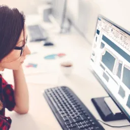 Krawcowa z ciemnymi włosami w okularach, pochylona nad monitorem z projektem szablonu odzieży, na biurku klawiatura i filiżanka kawy.