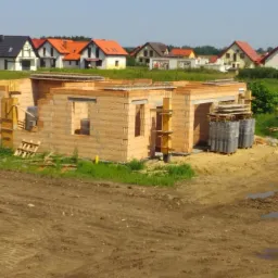 Wznoszenie ścian parterowego domu z bloczków ceramicznych na tle zabudowy jednorodzinnej i zieleni.