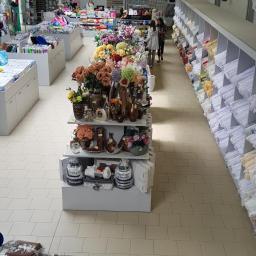 Prima Sp z Oo - Wnętrze sklepu z dekoracjami, ekspozycja sztucznych kwiatów w wazonach i akcesoriów dekoracyjnych na regałach, w tle regały z tkaninami i osoby robiące zakupy.