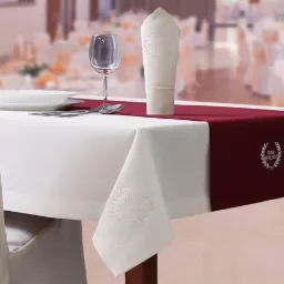 Elegancko nakryty stół w restauracji lub sali bankietowej, biały obrus z wytłoczonym logo, bordowy bieżnik, kieliszek do wina, talerz, sztućce, krzesło w pokrowcu z logo, w tle inne stoły...
