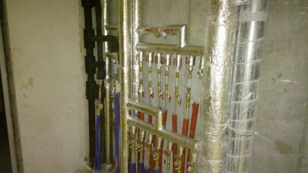 Rozdzielacz instalacji grzewczej z licznymi zaworami, rury izolowane folią aluminiową, widok na surową ścianę w tle.