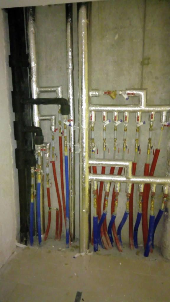 Rozdzielacz instalacji centralnego ogrzewania z rurami w izolacji, widoczne rury czerwone i niebieskie, oraz zawory z pokrętłami.