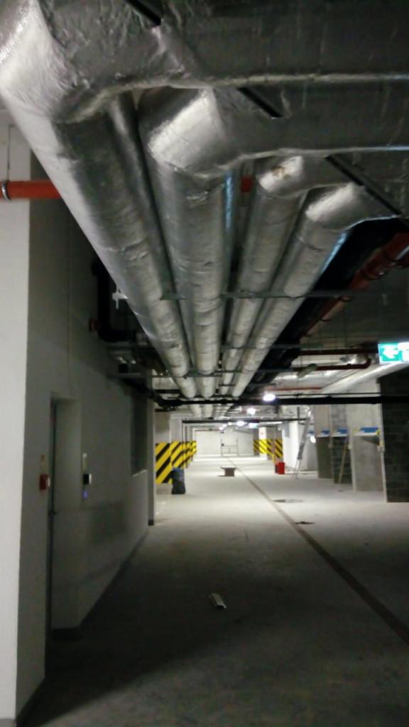 Instalacja wentylacyjna z izolacją termiczną, widoczna pod sufitem parkingu podziemnego, z rurami w srebrnej folii i elementami mocującymi, w tle kolumny z żółto-czarnymi pasami.