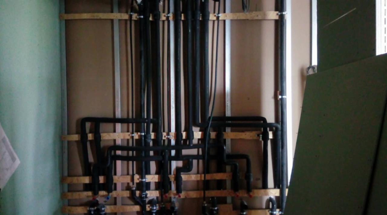 Czarne rury instalacji wodnej zamocowane do ściany za pomocą drewnianych wsporników i metalowych uchwytów, widoczne połączenia i izolacja.