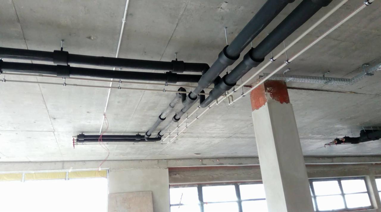 Czarno-szare rury instalacyjne podwieszone pod surowym betonowym sufitem w budynku w stanie deweloperskim, widoczne wsporniki i kable.