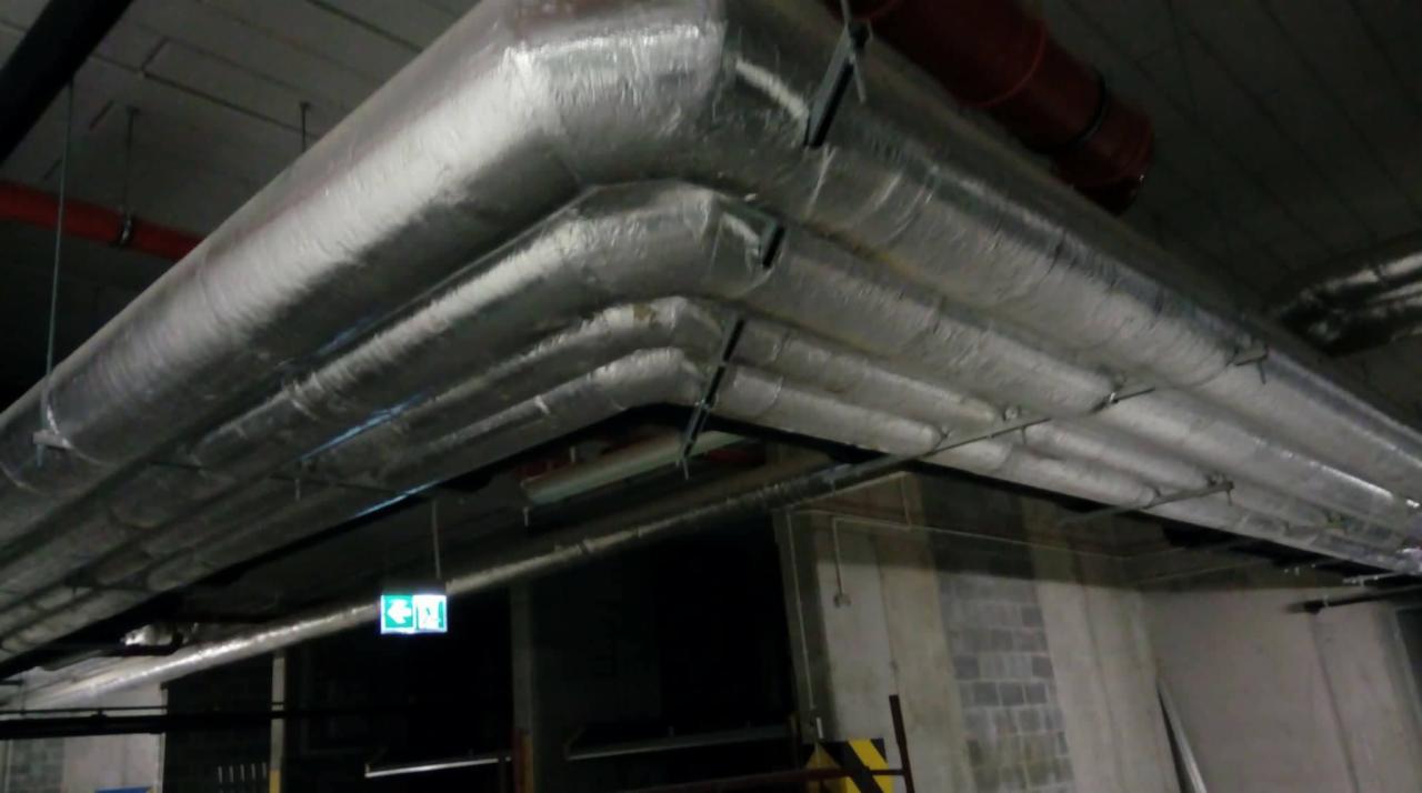 System srebrnych, izolowanych rur wentylacyjnych pod sufitem w hali przemysłowej, widoczny znak ewakuacyjny.
