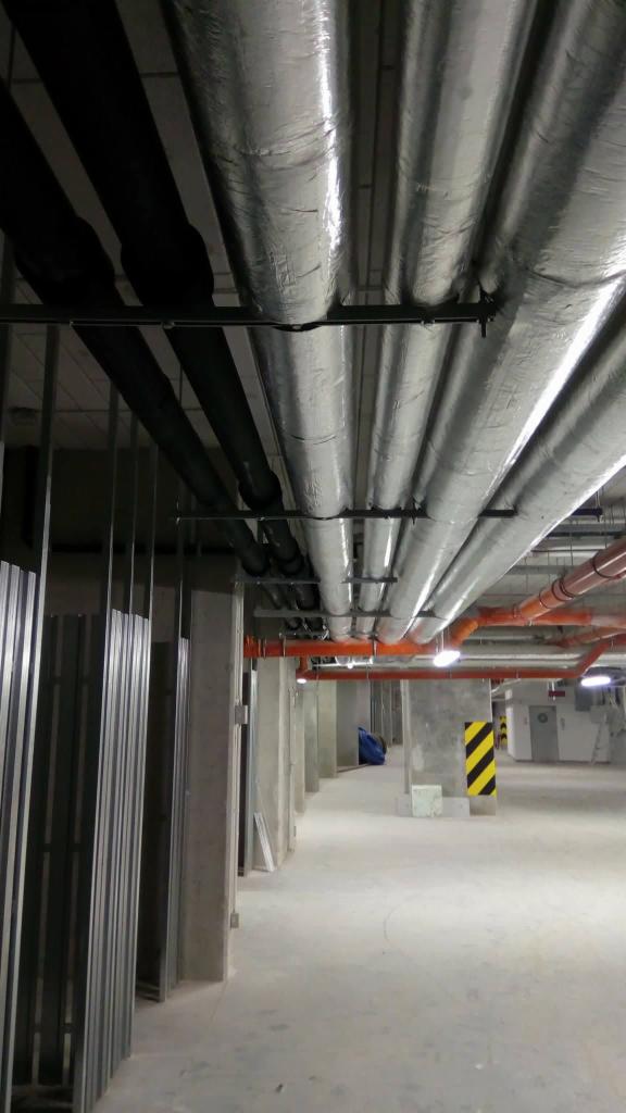 Rzędy rur instalacyjnych, niektóre owinięte folią aluminiową, biegnące pod sufitem parkingu podziemnego lub hali, wsparte na metalowych konstrukcjach.